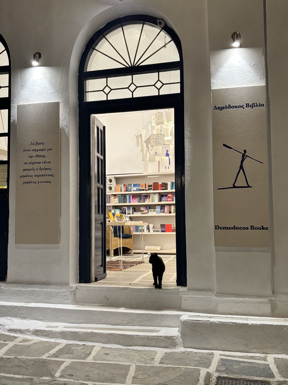 The lit entrance of Demodocos Books at dusk, Ios — the oar-bearer sign beside the door and the shop cat on the threshold